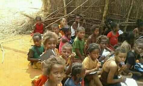 Photo of approximately 20 children in SW Madagascar sitting on the dirt ground with pencils and notebooks